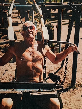 An elderly man with sunglasses is seen working out on a chest press machine outdoors in natural lighting, with foliage in the background.