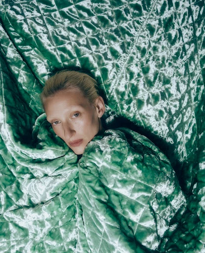 Adult fashion model lies on a green quilted fabric, posing with head tilted back looking toward the camera in indoor lighting.