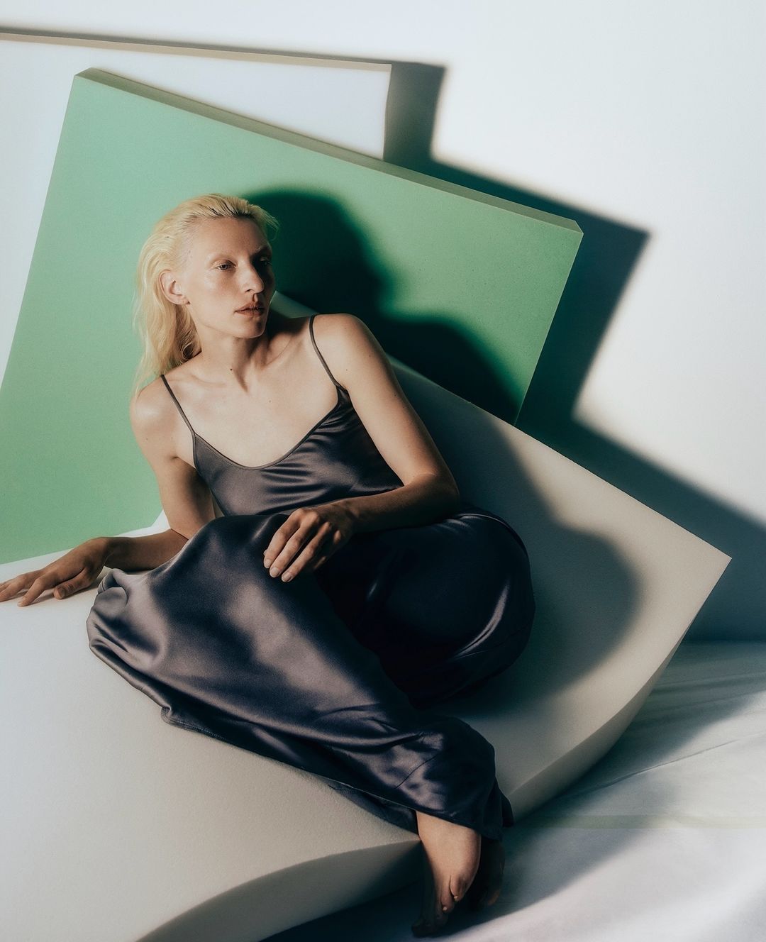 Woman in a long slip dress poses against geometric shapes in a studio. She has short blond hair and is barefoot.