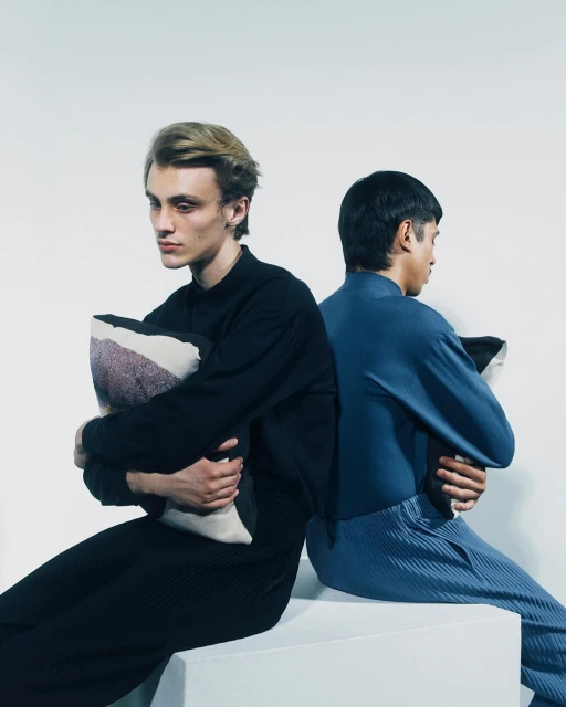 Two stylish men sit back-to-back in a photo studio, each holding a pillow. The man on the left wears black, and the man on the right wears blue.