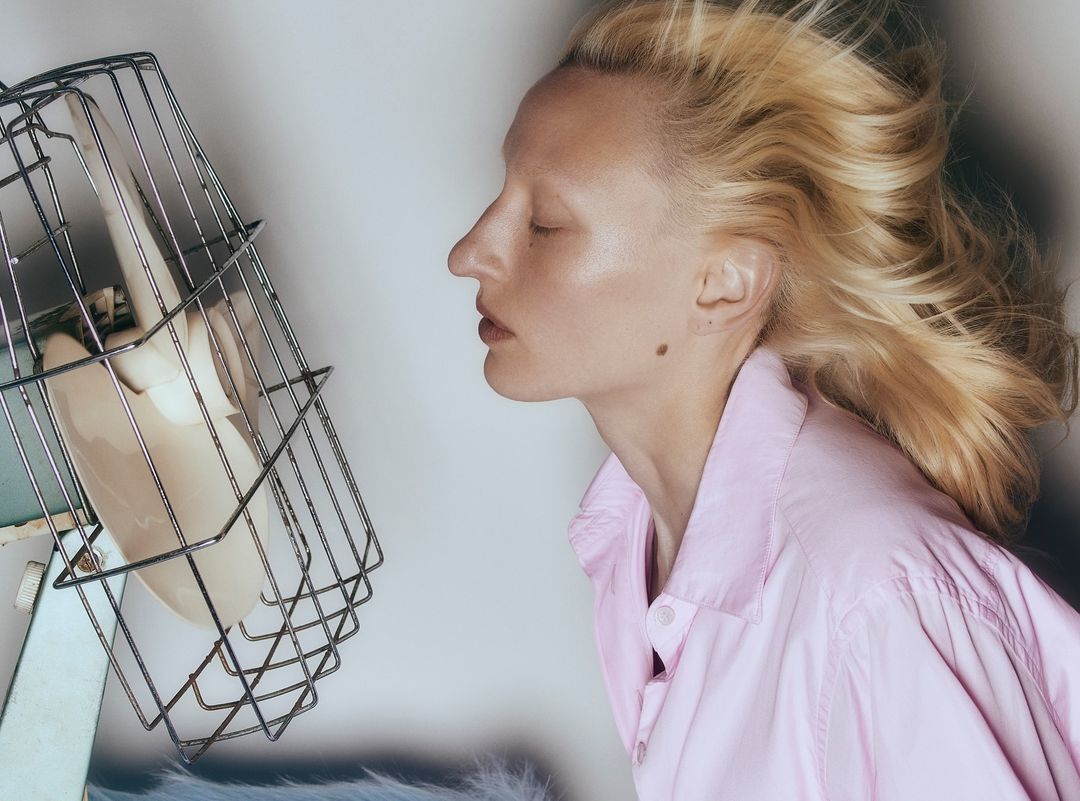 Woman in profile wearing a pink shirt with blonde hair blowing in the breeze of an electric fan indoors against a white background.