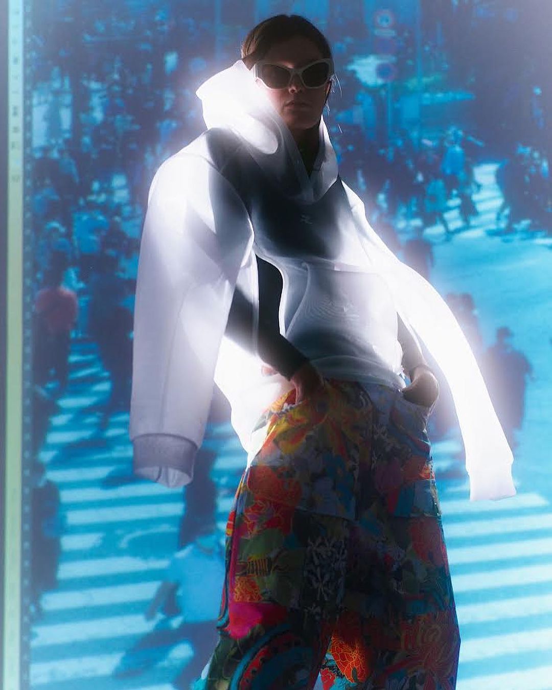 Woman poses in transparent hooded dress with crowd projection background, wearing sunglasses and colorful pants, in a vibrant fashion shot.
