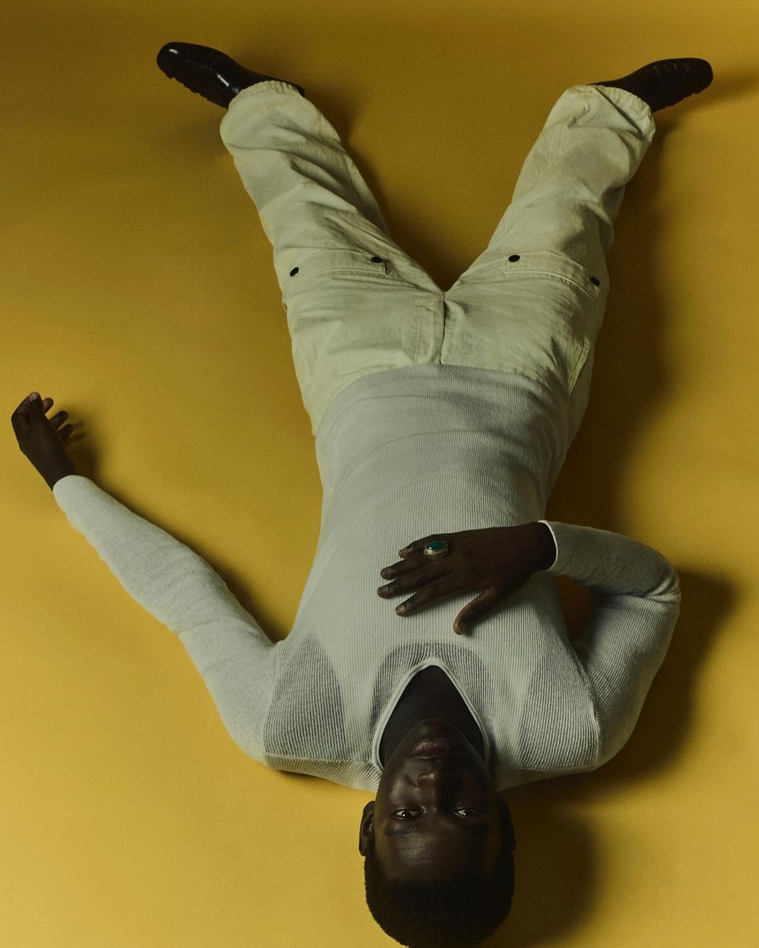 A dark-skinned adult man is lying down on a yellow surface wearing cream-colored pants, a sweater, and dark shoes.
