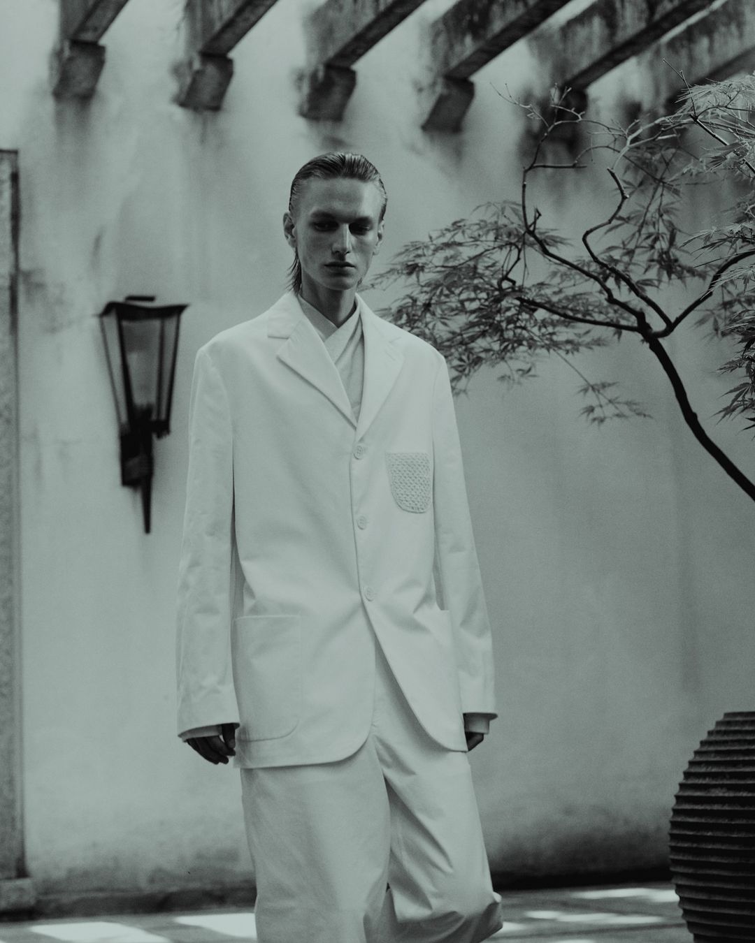 Monochromatic portrait of a slender male model in a white suit posing outdoors against a textured wall and decorative tree.