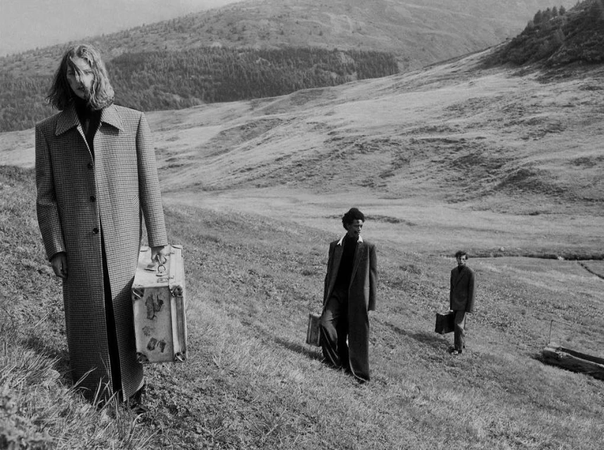 Three people with suitcases stand on a grassy hillside in a black and white image, with a mountain range in the background, evoking a sense of travel.