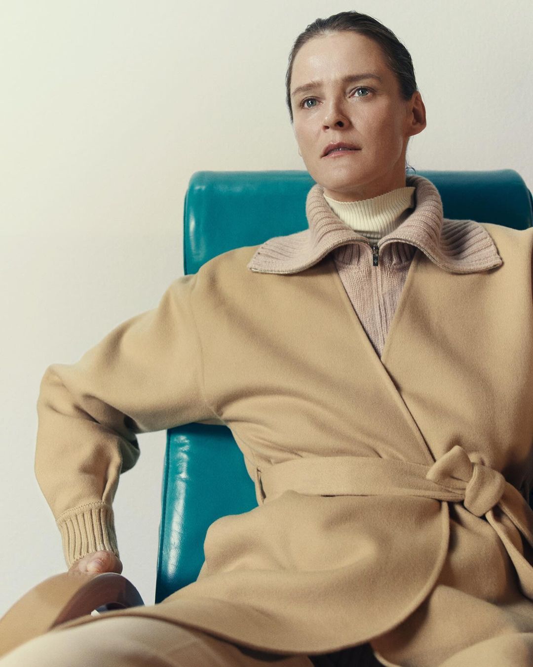 Adult woman in a tan coat sitting on a blue chair. She wears a light-colored sweater. The background is plain and bright. The photo is indoors.