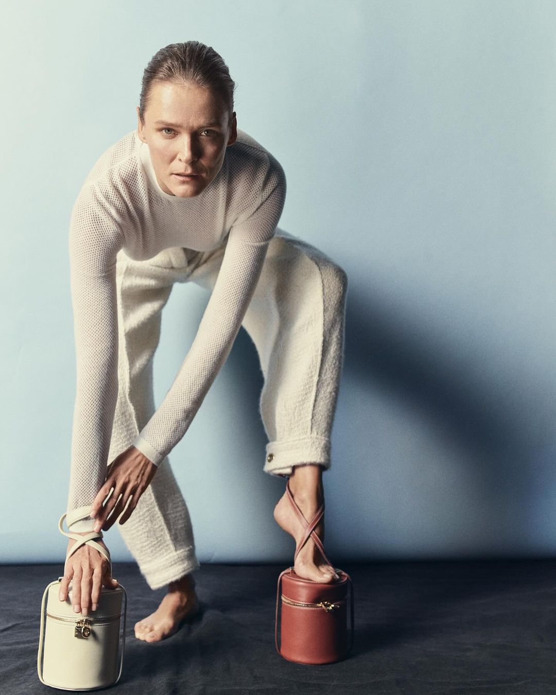 Woman in white outfit poses barefoot with cream and brown handbags against a blue background in a fashion editorial shot.