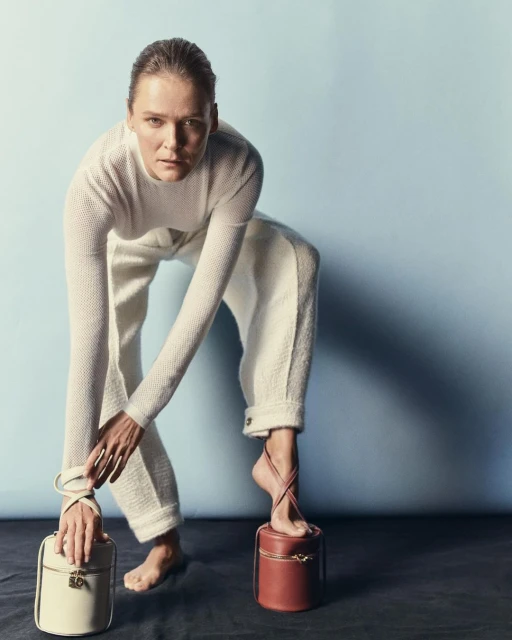 Woman in white outfit poses barefoot with cream and brown handbags against a blue background in a fashion editorial shot.