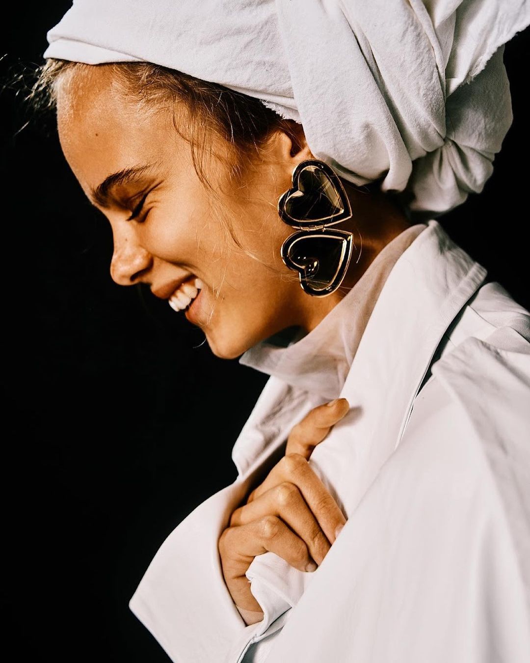 Woman smiles in white shirt and headscarf with large gold heart earrings against a dark background in a fashion-forward portrait.