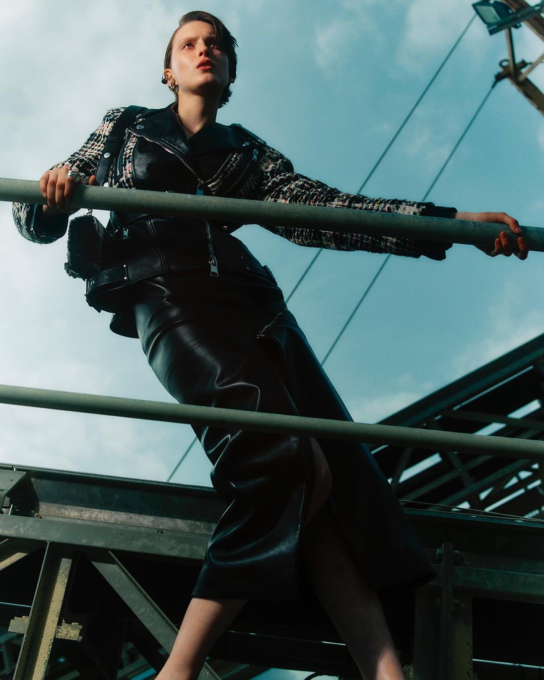 Fashion shot of a woman posing on a metal structure against a blue sky, wearing a leather skirt and jacket, and a patterned blazer.