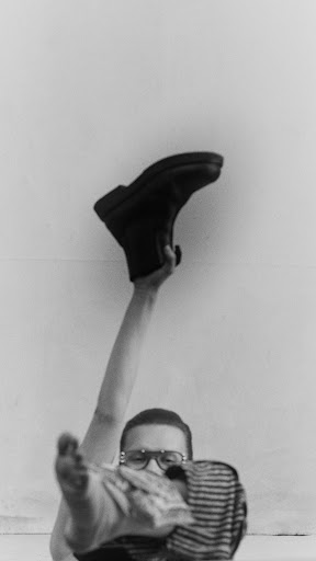 Monochrome photo of a person in glasses holding up a black boot against a white wall, capturing a minimalist and stark visual.