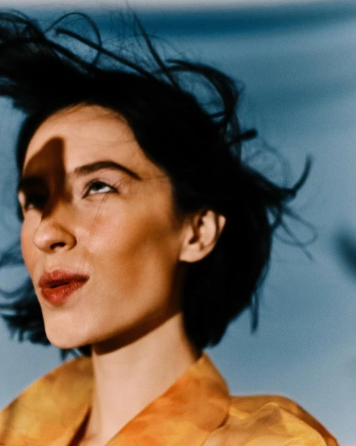 Portrait of an adult woman with dark hair, wearing an orange patterned shirt, gazing upwards against a soft blue sky backdrop.