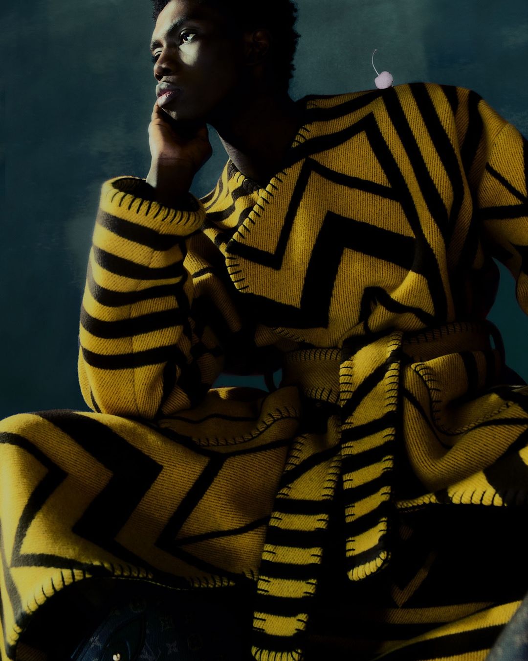 An adult man poses indoors, wearing a yellow and black zigzag-striped robe, looking off to the side in a portrait-style fashion shot.