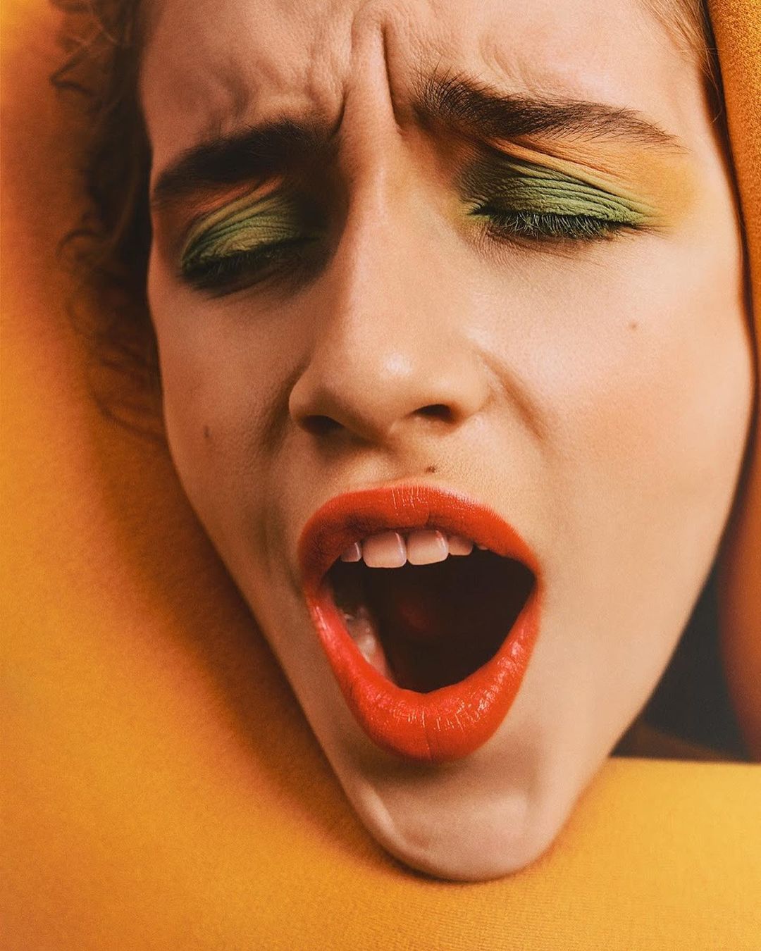 Close-up portrait of a woman with vibrant green eyeshadow and bright red lipstick, her mouth open in a dynamic expression.