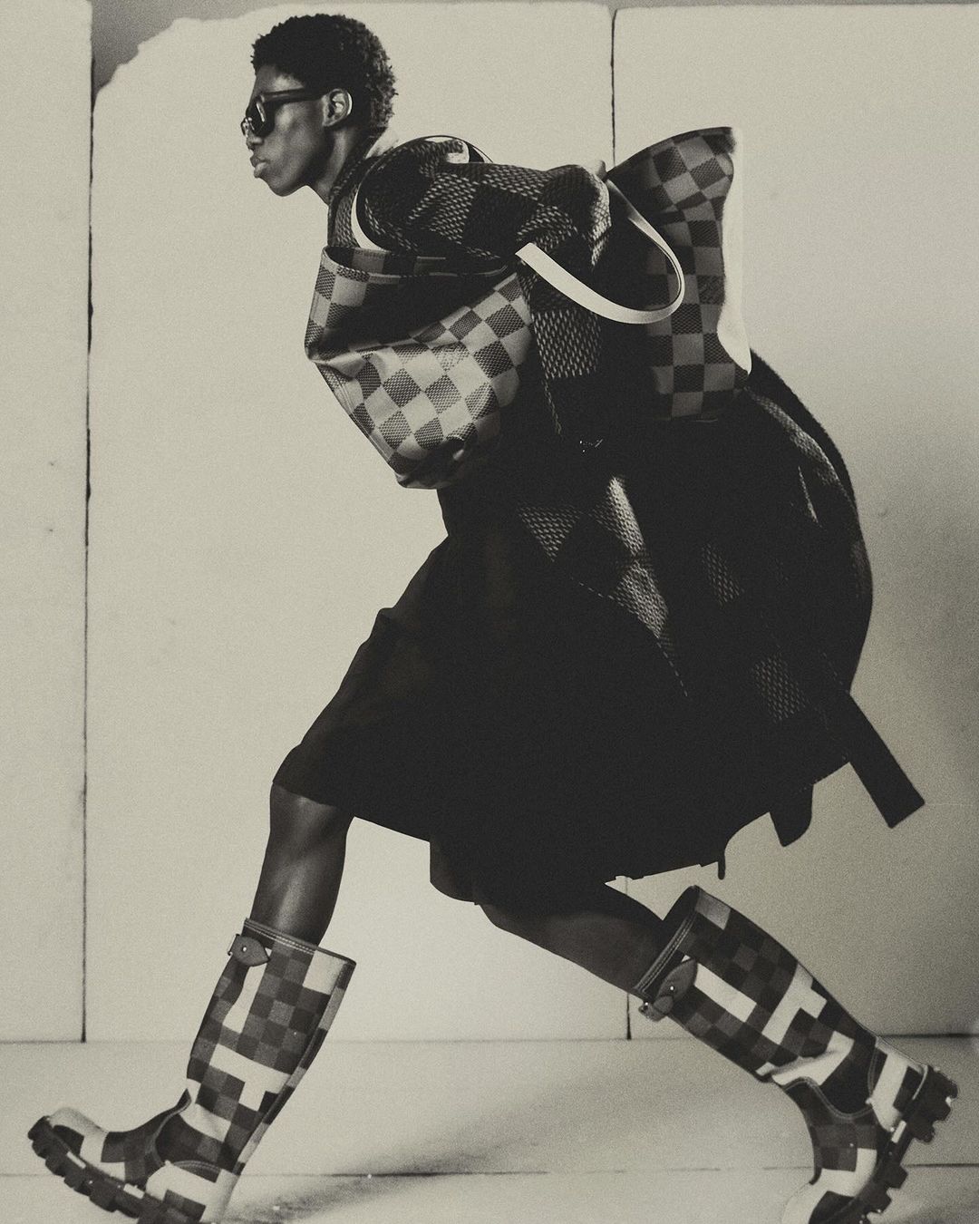 Fashion editorial shot of a Black male model in checkered rain boots, skirt, jacket, and matching bag, posing in a photo studio.