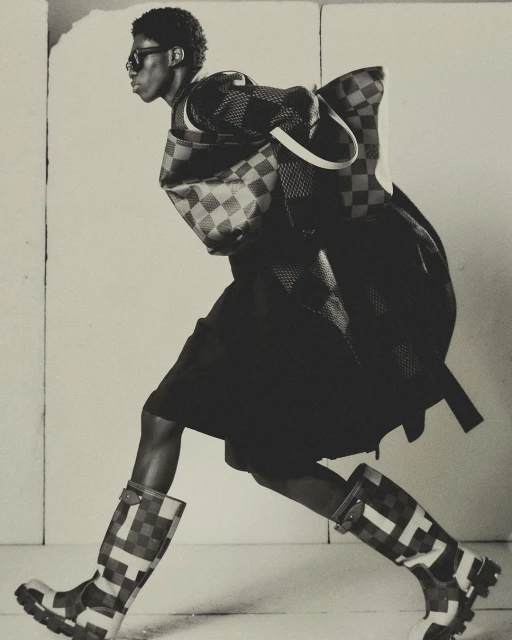 Fashion editorial shot of a Black male model in checkered rain boots, skirt, jacket, and matching bag, posing in a photo studio.