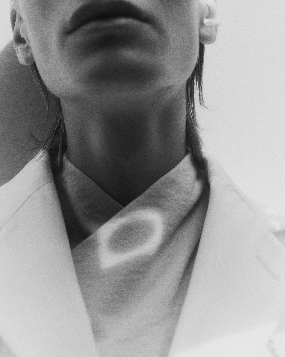 Close-up black and white shot of a person's face, neck, and shoulders wearing a white coat, showcasing a minimalist and modern aesthetic.