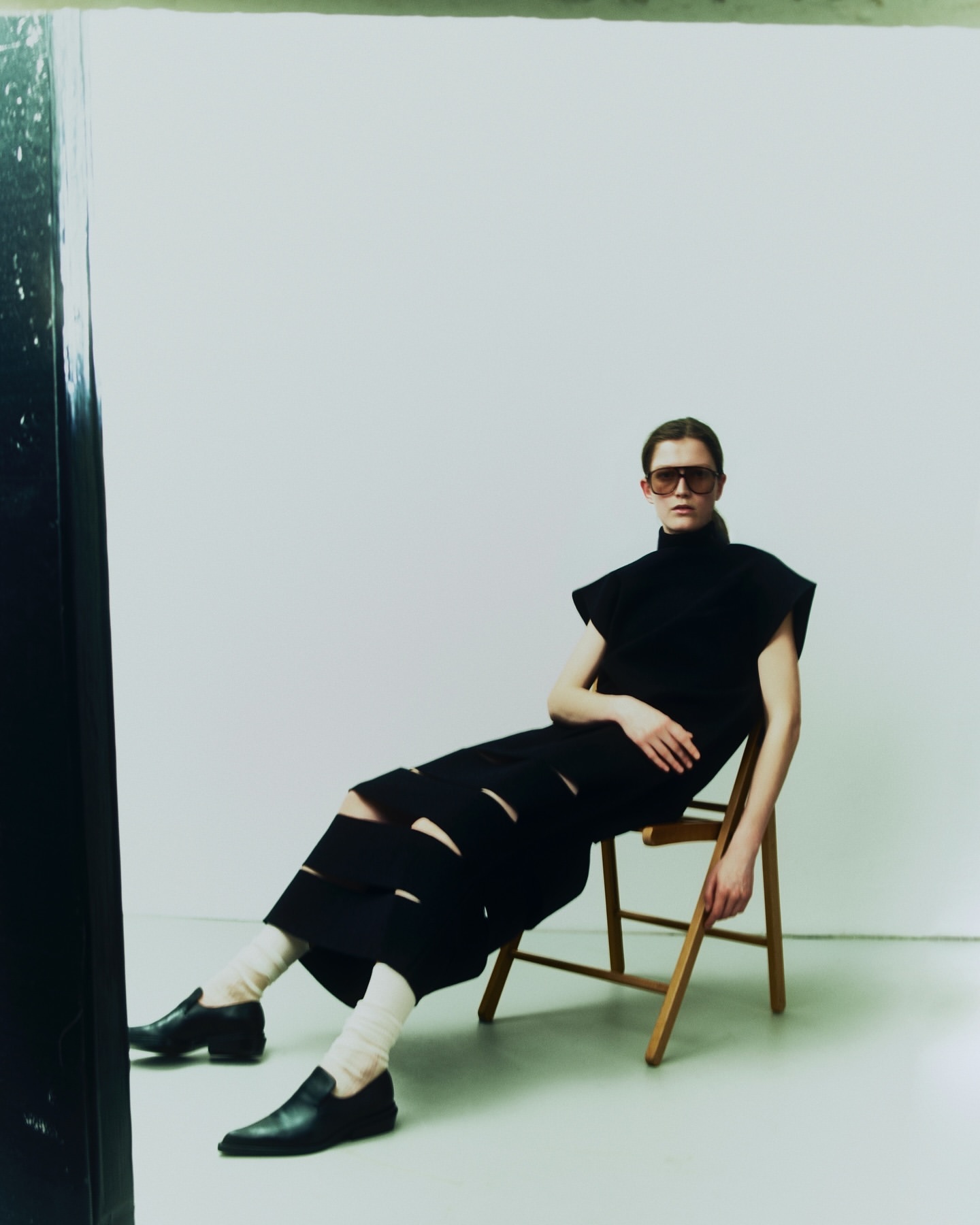 A model in a black outfit with cutouts poses while sitting on a wooden chair against a white backdrop inside a photo studio.