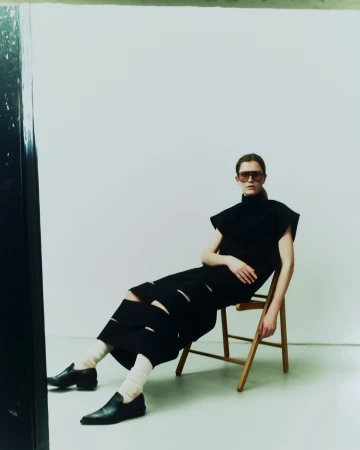 Model wearing dark sunglasses and a black cut-out dress seated on a wooden folding chair against a plain white wall