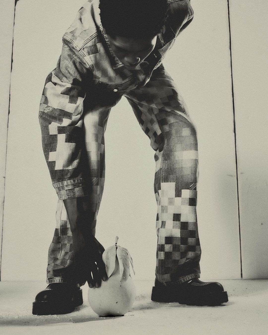 Person in patterned denim poses with a pear in a minimalist photo studio. Black and white fashion portrait.