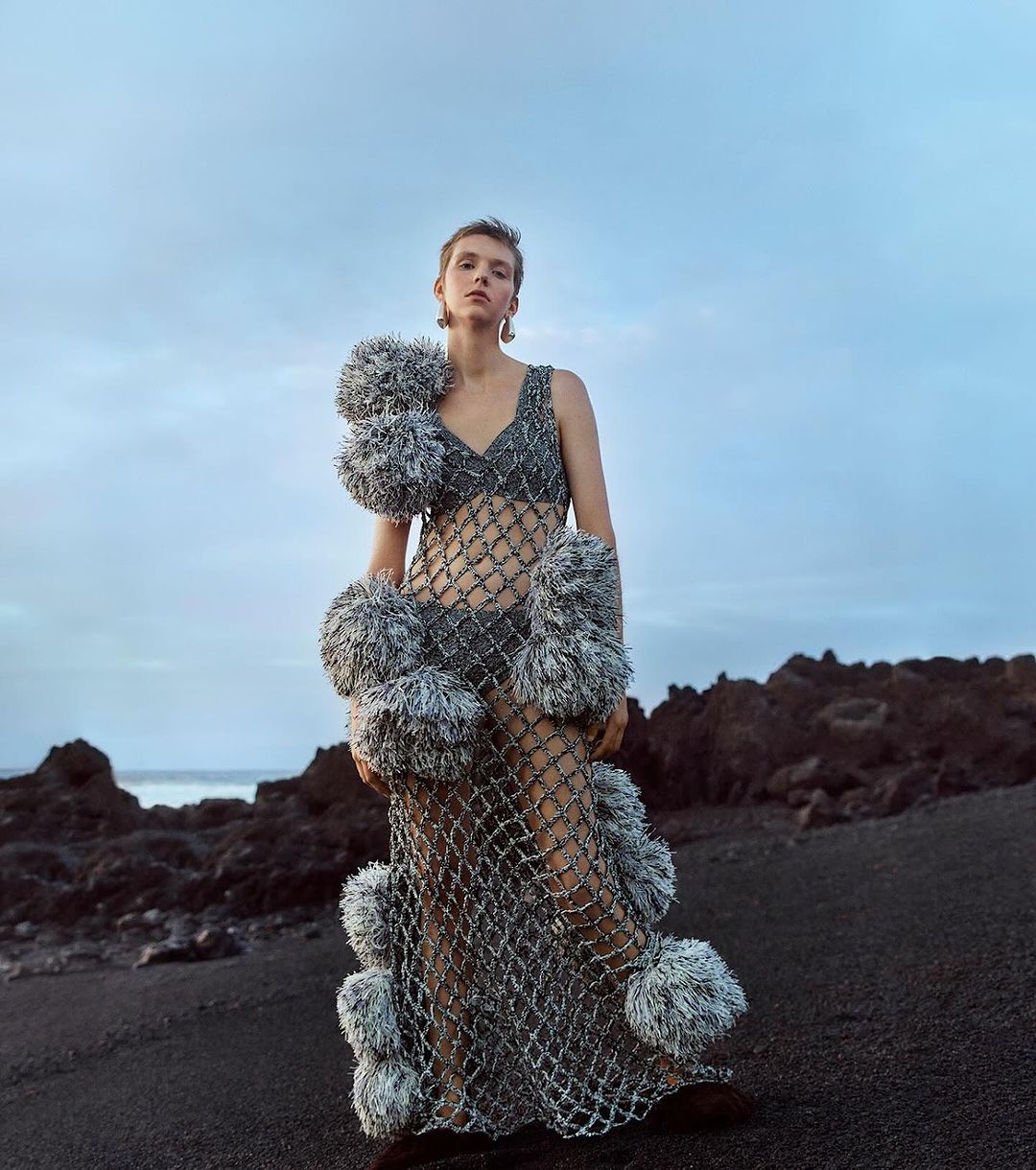 A woman in a mesh dress with pom-poms poses on a black sand beach with a cloudy sky and volcanic rocks in the background, fashion.