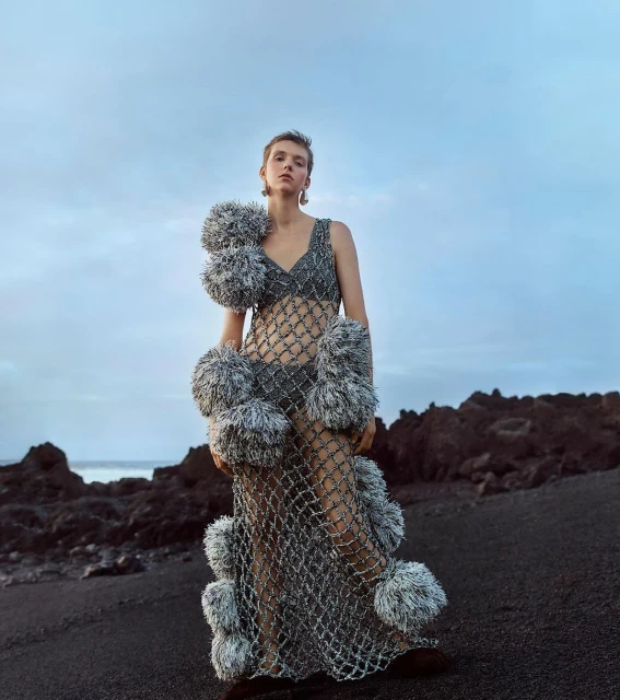 A woman in a mesh dress with pom-poms poses on a black sand beach with a cloudy sky and volcanic rocks in the background, fashion.