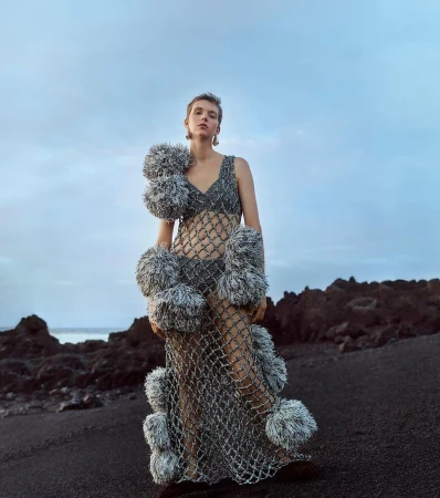 A woman in a mesh dress with pom-poms poses on a black sand beach with a cloudy sky and volcanic rocks in the background, fashion.