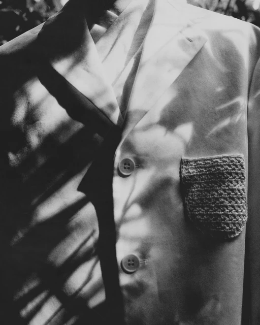 Close-up of a light-colored suit jacket with a knitted pocket detail, showcasing texture and tailoring under soft, dappled lighting.