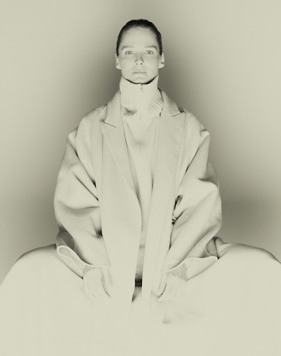 Woman wearing a light-colored coat and turtleneck sweater sitting in front of a white background in a fashion photography studio shot.