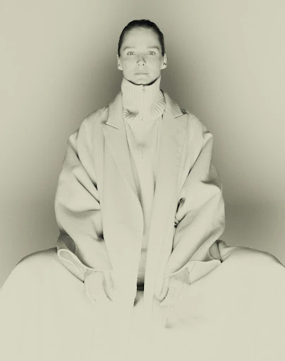 Woman wearing a light-colored coat and turtleneck sweater sitting in front of a white background in a fashion photography studio shot.