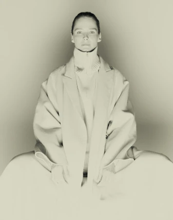 Monochrome studio portrait of a woman wearing an oversized coat and turtleneck sweater against a plain background.