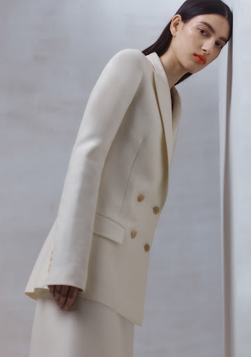 Elegant woman in cream-colored double-breasted blazer and matching skirt, leaning against a wall in a neutral, minimalist indoor setting.