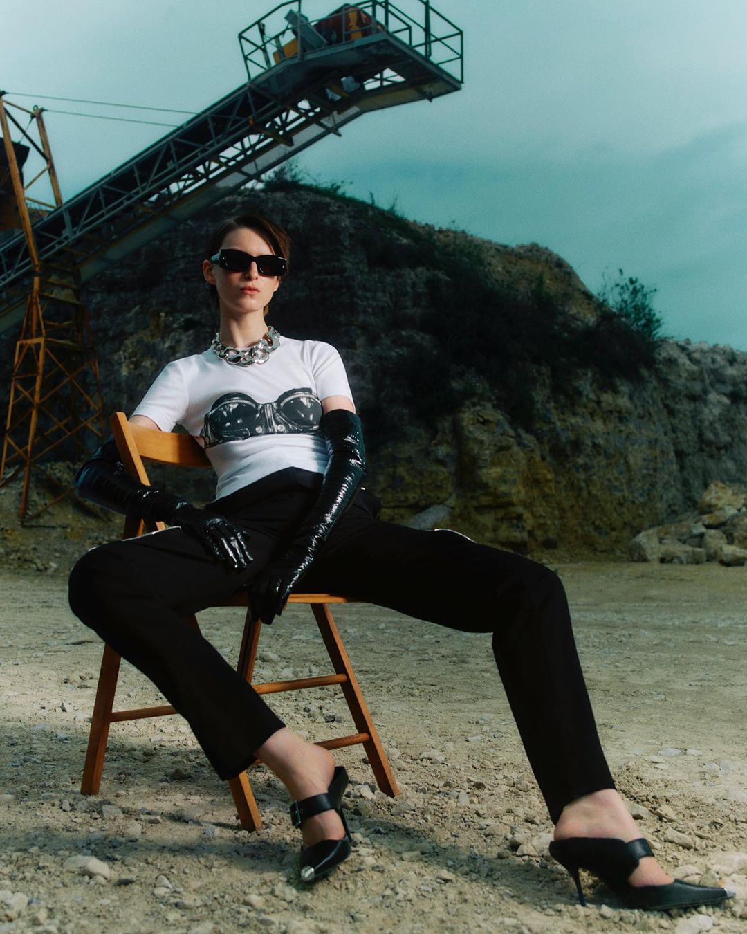 Elegant adult female model in black gloves, sunglasses, and heels, sitting on a chair against an industrial quarry backdrop.