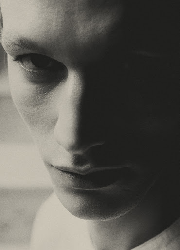 Close-up studio portrait of a young adult man with a serious expression, lit with dramatic chiaroscuro lighting, black and white photography.