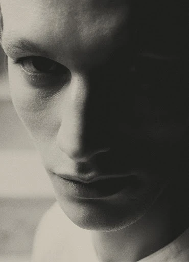 Close-up studio portrait of a young adult man with a serious expression, lit with dramatic chiaroscuro lighting, black and white photography.