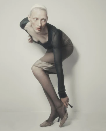 Model in a black top and skirt adjusting a high-heel shoe against a plain light grey background