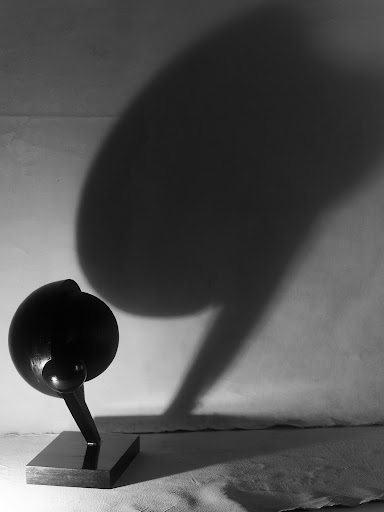 Abstract art sculpture with a round shape on a stand, creating a large shadow against a neutral wall in a monochromatic composition.