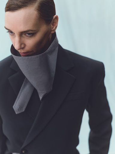 An adult female model in a grey neckpiece and a black blazer poses against a light blue background in a fashion editorial studio portrait.