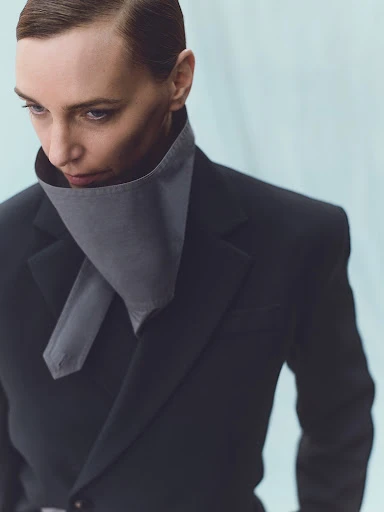 An adult female model in a grey neckpiece and a black blazer poses against a light blue background in a fashion editorial studio portrait.