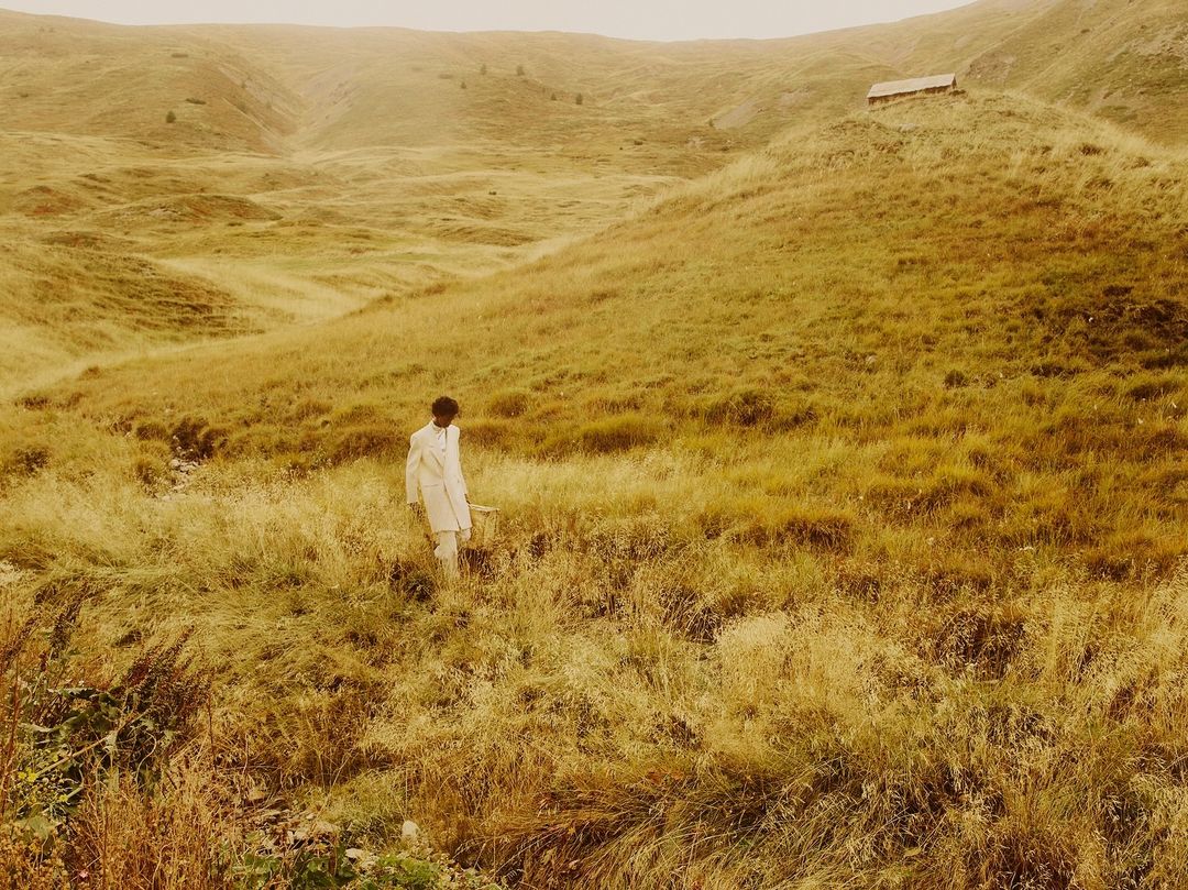 Man wearing a white coat walking through a grassy meadow by Nicolas Kern