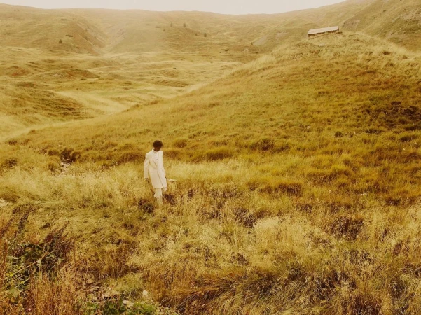 Man wearing a white coat walking through a grassy meadow by Nicolas Kern