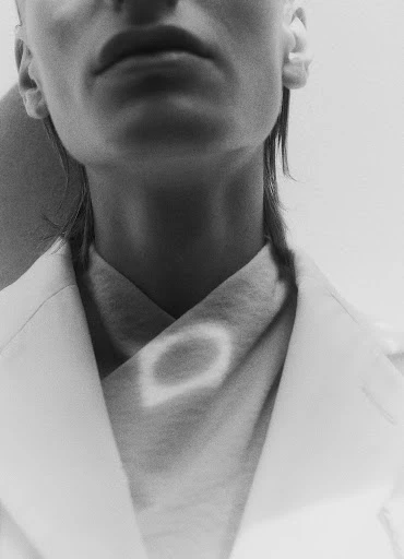 Monochrome close-up of an adult with short hair wearing a light colored jacket and scarf against a light background in a studio.