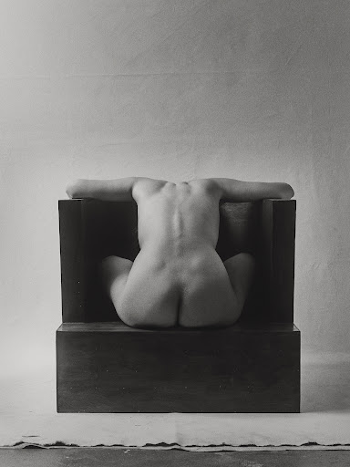 Monochrome nude art photo of a seated, back-facing adult female with arms outstretched, positioned against a dark block and soft backdrop.