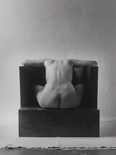 Monochrome nude art photo of a seated, back-facing adult female with arms outstretched, positioned against a dark block and soft backdrop.