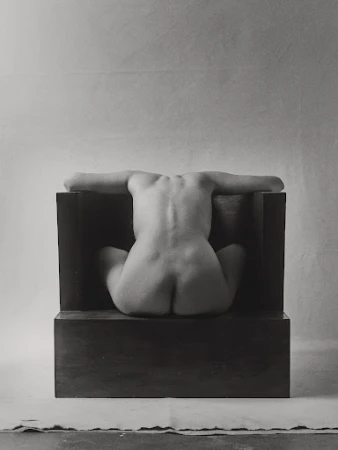 Monochrome nude art photo of a seated, back-facing adult female with arms outstretched, positioned against a dark block and soft backdrop.