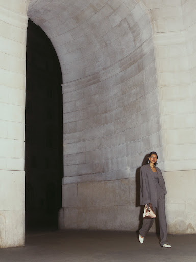 A woman in a suit walks through a stone archway, holding a purse, with neutral tones highlighting the architecture and fashion.