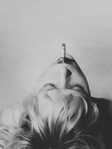 Black and white close-up of a woman with closed eyes, laying down with a cigarette in her mouth, against a plain backdrop.