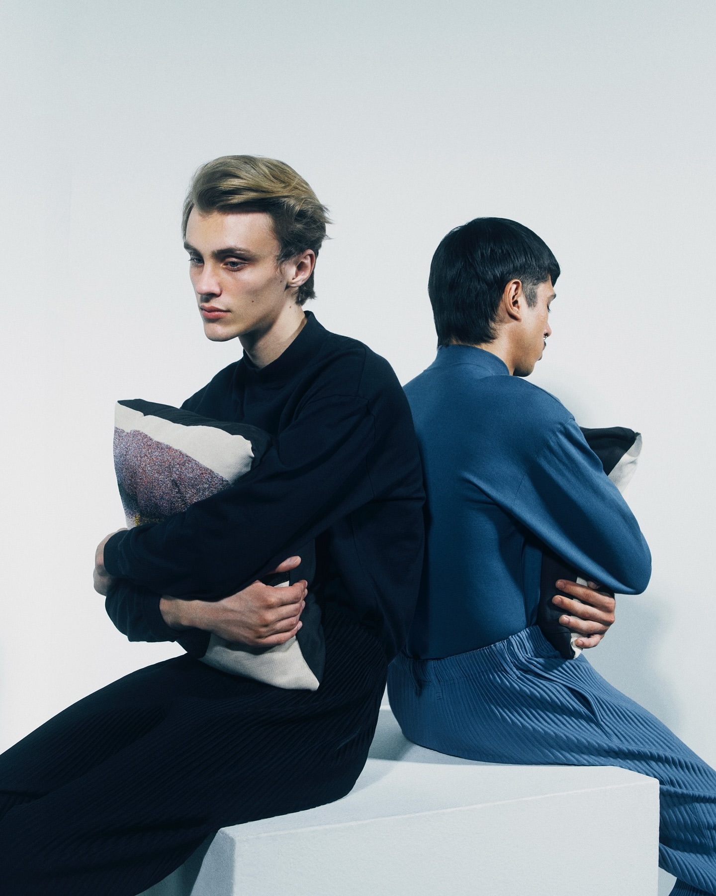 Two young adults sit back-to-back on a white block, holding pillows, set against a plain white backdrop in a studio environment.