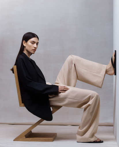 Model wearing a black blazer and beige trousers seated on a wooden zig zag chair with her foot pressed against a white wall.