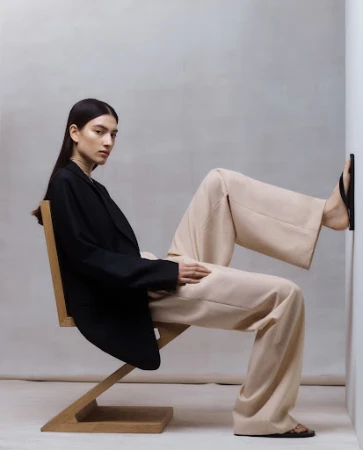 Model wearing a black blazer and beige trousers seated on a wooden zig zag chair with her foot pressed against a white wall.